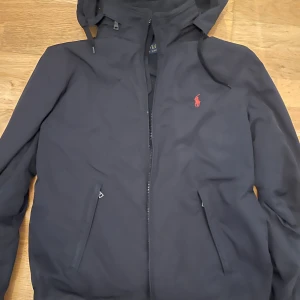 Svart vindjacka från Polo Ralph Lauren - Svart vindjacka från Polo Ralph Lauren med röd logga på bröstet. Jackan har huva, dragkedja framtill och två snedställda fickor med dragkedja. Insidan är fodrad med mjuk fleece för extra komfort. Perfekt för kyliga dagar.