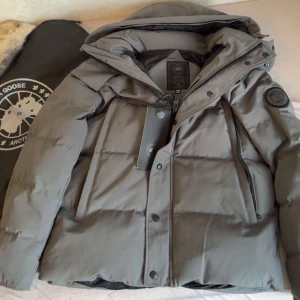 Grå pufferjacka från Canada Goose - Säljer en grå pufferjacka från Canada Goose med huva och snyggt broderat logomärke på ärmen. Jackan har dragkedja och knappar framtill, samt praktiska fickor med dragkedja. Perfekt för kalla dagar och riktigt stilren look.Storlek M