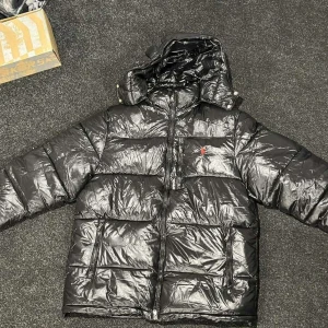 Svart pufferjacka från Polo Ralph Lauren - Säljer en svart, glansig pufferjacka från Polo Ralph Lauren med röd broderad logga på bröstet. Jackan har huva, dragkedja framtill och två stora fickor med dragkedja. Perfekt för kalla dagar och riktigt snygg streetstil.