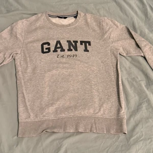 Grå sweatshirt från GANT med tryck - Snygg grå sweatshirt från GANT med stort svart tryck på bröstet och texten 'Est. 1949'. Tröjan har rund hals, ribbade muddar och lång ärm. Insidan är mjuk och skön, perfekt för chill dagar. Klassisk och enkel design med diskreta detaljer.