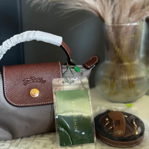 Väska  - Snygg handväsk i brun/grått med bruna detaljer i skinn. Väskan har guldiga metalldetaljer, klassisk knappstängning och präglad logotyp på locket. Kommer med axelrem och påse. Perfekt storlek för det viktigaste. Skriv privat för fler frågor och innan du beställer. 💕