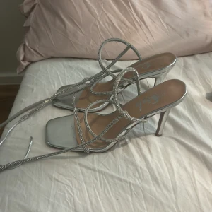 Glittriga silver sandaler med klack - Säljer ett par snygga sandaler i silver med glittriga remmar och hög klack. Skorna har fyrkantig tå och tunna remmar som går runt foten och ankeln. Perfekta för att sticka ut med en glamorös vibe. Ett band saknas