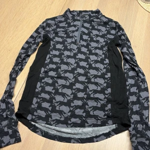 Svart och grå långärmad tröja CRW - Cool långärmad tröja från CRW i svart och grått med hästmönster över hela plagget. Tröjan har en half zip framtill och är tillverkad i mjukt bambumaterial. Perfekt för dig som gillar ridning och vill ha en unik look.