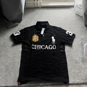  pikétröja Ralph Lauren Chicago Chief Keef - Svart pikétröja från Ralph Lauren med broderad logga, stort CHICAGO-tryck och guldfärgad emblem på bröstet. Nummer 3 på ärmen och ryggen. Slim fit modell med korta ärmar och klassisk krage. Perfekt för dig som gillar sportig och stilren look.
