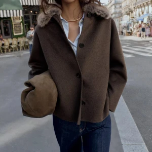 Brun ulljacka med fuskpäls-krage - Snygg brun jacka i ull med rak passform och stora svarta knappar framtill. Kragen är klädd i mjuk fuskpäls i beige nyans som ger en cool kontrast. Jackan har lång ärm och en minimalistisk look som passar perfekt till jeans.