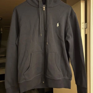 Mörkblå hoodie från Polo Ralph Lauren - Snygg mörkblå hoodie från Polo Ralph Lauren med klassisk logga broderad på bröstet. Tröjan har huva med snörning, dragkedja framtill och två stora fickor. Perfekt för chill dagar och har en clean, stilren look.