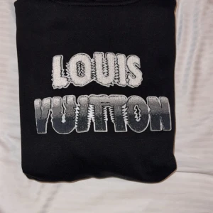 Svart Louis Vuitton hoodie S - Svart hoodie från Louis Vuitton med stor broderad logga i vitt och grått på bröstet. Tröjan har huva med snörning, känguruficka och en orange patch med logga på ryggen. Materialet är mjukt och känns som bomull, perfekt för dig som gillar exklusiva streetwear-vibes.