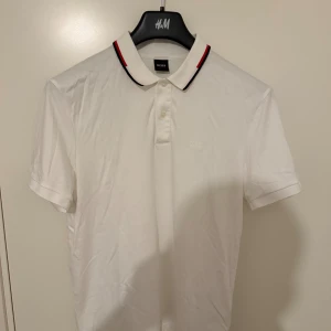 Vit piké från BOSS med röd detalj - Vit pikétröja från BOSS med klassisk krage och knappar framtill. Kragen har en snygg röd och svart rand som ger en sportig touch. Tröjan är kortärmad och tillverkad i mjuk bomull för en skön känsla.