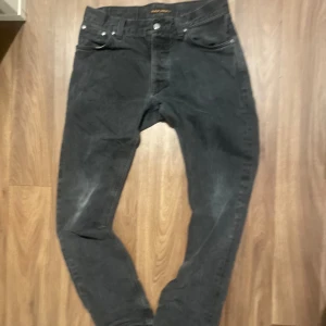 Svarta jeans från Nudie Jeans W31 L36 - Svarta jeans från Nudie Jeans i storlek W31 L36, tillverkade av 100% ekologisk bomull. Klassisk femficksmodell med svarta sömmar och subtila slitningar. Snyggt broderade bakfickor och knappgylf. Perfekt för dig som gillar stilrena och hållbara jeans.