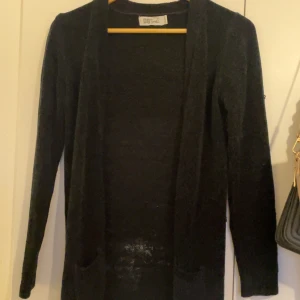 Svart lång kofta från DB Soul - Svart lång kofta från DB Soul med öppen front och två fickor framtill. Koftan är stickad och har långa ärmar, perfekt att slänga över en t-shirt eller topp. Enkel och stilren design som funkar till många outfits.