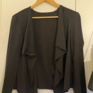 Svart omlott cardigan jacka - Svart cardigan jacka med omlott-design och långa ärmar. Jackan har en mjuk och flowy look framtill med öppet fall, perfekt att slänga över en t-shirt eller topp. Enkel och stilren, passar dig som gillar minimalistisk stil.