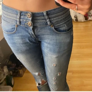 Lågmidjade bootcut jeans med slitningar - Snygga blå lågmidjade jeans med bootcut och slitna detaljer på benen. Modellen har låg midja med dubbla knappar och klassisk femficksdesign. Jeansen har en cool tvätt och slitningar som ger en edgy vibe. Perfekt för dig som gillar en avslappnad och trendig stil. Midjemåttet rakt över är 38cm och innerbenslängden är 80cm. Står storlek 26💗märket är fishbone. Har sytt om till bootcut själv