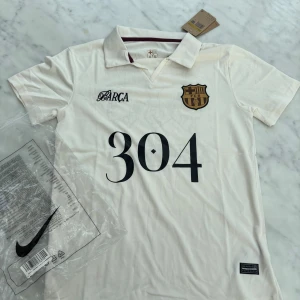 Vit FC Barcelona fotbollströja Nike S - Säljer en vit FC Barcelona fotbollströja från Nike i storlek S. Tröjan har korta ärmar, krage och tryck med '304' på framsidan samt klubbmärke. Baksidan har ett stort motiv med Lamine Yamal och Barça-logga. Materialet är lätt och andas, perfekt för match eller träning.