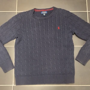Polo Ralph Lauren stickad tröja - Säljer en mörkblå stickad tröja från Polo Ralph Lauren i klassisk kabelstickad design. Tröjan har rund halsringning, långa ärmar och en röd broderad logga på bröstet. Perfekt för dig som gillar stilrena och tidlösa plagg. Bra skick. Hör av er vid minsta fundering❗️Skynda er det är väldigt bra pris!