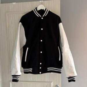 Baseballjacka svart och vit - Säljer en klassisk baseballjacka i svart med vita ärmar och vita detaljer av äkta läder. Jackan har ribbade muddar och krage med marinblå och vita ränder, knäppning med vita knappar och två fickor fram. Fodret är quiltat och insidan har en snygg etikett. Perfekt för en sportig och cool look. Pris kan diskuteras, hör av dig vid minsta fråga!