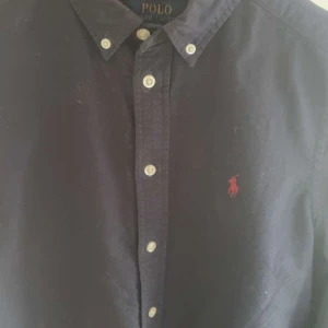Mörkblå skjorta från Polo Ralph Lauren - Säljer en klassisk mörkblå skjorta från Polo Ralph Lauren med knappar framtill och röd broderad logga på bröstet. Skjortan har en tidlös design med button-down krage och är perfekt för dig som gillar stilrena plagg.