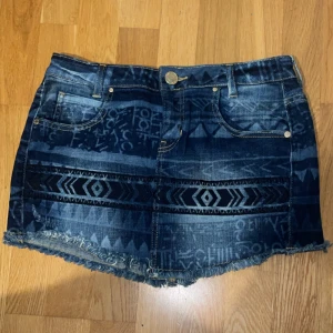 Mönstrad jeanskjol från Guess - STORLEK 27. Snygg kort jeanskjol från Guess med coolt mönster i olika blå toner och slitna kanter. Kjolen har klassiska fickor fram och bak, bälteshällor och knappstängning. Perfekt för dig som vill sticka ut med en unik denimlook.
