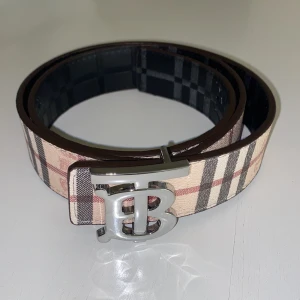 Burberry bälte - Snyggt bälte från Burberry med deras ikoniska rutiga mönster i beige, svart och rött. Bältet har ett stort silvrigt spänne med Burberrys. Bra kvalité!.