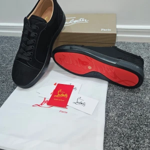 Svarta sneakers från Christian Louboutin - Snygga svarta sneakers från Christian Louboutin med klassisk röd sula och diskret logga på sidan. Skorna har rund tå, platt sula och är tillverkade i mocka med svarta snören. Insidan är beige och har guldfärgad logotyp. Perfekt för dig som gillar exklusiv streetstyle.