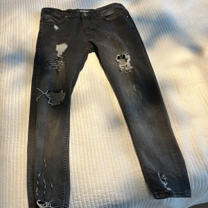 Svarta skinny jeans med slitningar - Från Paris lokalt märke .Svarta skinny jeans med kraftiga slitningar och hål på benen samt tryckt textdetalj på vänster lår. Jeansen har en smal passform och klassisk femficksdesign. Materialet är denim och färgen är tvättad svart med ljusare partier.