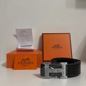 Svart H skärp med H-spänne - Exklusivt svart skärp från H med klassiskt H-spänne i silverfärgad metall. Bältet är tillverkat i äkta skinn med tydlig struktur och har vita kontrastsömmar längs kanterna. Perfekt accessoar för dig som gillar lyxiga detaljer och stilren design.