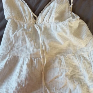 Vit kort klänning med spetsdetaljer - Supersöt vit kort klänning med tunna axelband och v-ringad hals med spetskant. Klänningen har en luftig, rynkad struktur och knytband framtill. Perfekt för varma dagar och har en lätt, flowy känsla.