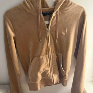 Beige velour hoodie från Juicy Couture - Säljer en beige hoodie från Juicy Couture i mjuk velour med dragkedja framtill, huva med snörning och känguruficka. Ikonisk JC-logga broderad på bröstet. Perfekt för en chill och trendig look.