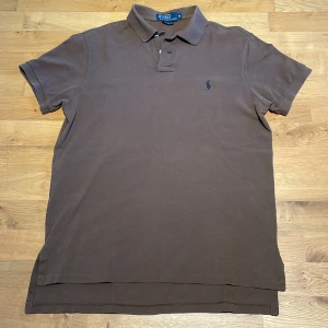 Brun pikétröja från Ralph Lauren M - Säljer en brun pikétröja från Polo Ralph Lauren i storlek M. Klassisk krage, kort ärm och broderad logga på bröstet. Tröjan är i bomull och har en snygg, enkel design som funkar till det mesta. Custom fit-modell. Den är längre baktill. Storlek: M.