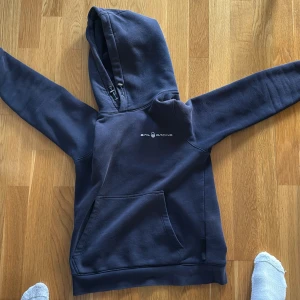 Mörkblå hoodie från Sail Racing - Mörkblå hoodie från Sail Racing med vit logga på bröstet. Tröjan har huva med snörning och en stor magficka framtill. Skön och avslappnad passform, perfekt för kyliga dagar. Materialet är mjukt och känns riktigt bekvämt.