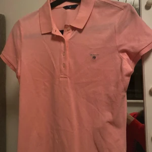 Rosa pikétröja från Gant - Säljer en klassisk rosa pikétröja från Gant med korta ärmar och krage. Tröjan har knäppning med fyra knappar framtill och Gant-logga broderad på bröstet. Perfekt för en sportig och stilren look. Kostade 300 kr 