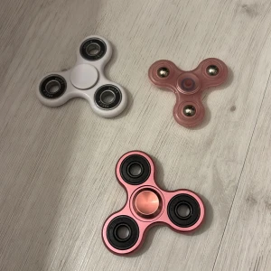 Fidget Spinner 3-pack - Säljer tre fidget spinners i olika färger: vit, rosa och metallic rosa. Alla fungerar utmärkt och spinner smidigt, vilket syns på bilderna. Inga synliga repor eller skador, i mycket gott skick. Perfekt för stresshantering eller som leksak.