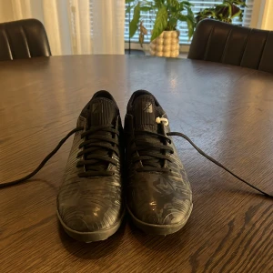 Svarta fotbollsskor från Puma - Säljer ett par svarta fotbollsskor från Puma med coolt kamouflagemönster och snörning. Skorna har rund tå, platt sula med dobbar och ovandel i syntetmaterial. Perfekta för dig som vill ha stil på planen och gillar sportiga detaljer.