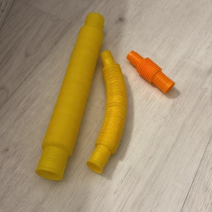Fidget/leksaks tubar - Bilderna visar färgglada plastleksaker, troligen s.k. pop tubes, som kan böjas och dras ut för att skapa olika former och ljud. De är i gult och orange och ser ut att vara i gott skick utan synligt slitage. Perfekt för sensorisk lek och kreativitet för barn.