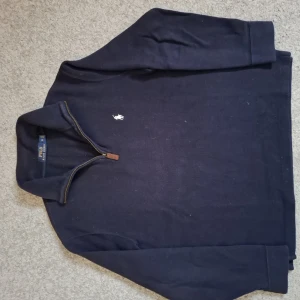 Mörkblå half zip tröja Polo Ralph Lauren - Nypris 2000 kr.  Mörkblå långärmad tröja från Polo Ralph Lauren med klassisk vit broderad logga på bröstet. Tröjan har en snygg half zip-dragkedja med läderdetalj och är tillverkad i mjuk bomull. Perfekt för en stilren och avslappnad look.