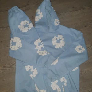 Blå hoodie från Denim Tears med blommor - Säljer en ljusblå hoodie från Denim Tears med vita blommiga tryck över hela tröjan. Hoodien har huva, känguruficka och ribbade muddar. Materialet är mjukt och fleecefodrat, perfekt för chill dagar. Passformen är loose och ärmarna är långärmade.