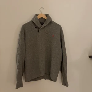 Grå tröja från Polo Ralph Lauren - Säljer en grå långärmad tröja från Polo Ralph Lauren med klassisk krage och två knappar vid halsen. Tröjan har det ikoniska röda broderade logot på bröstet och är tillverkad i mjuk bomull. Perfekt för lager-på-lager under höst och vinter. Om inte priset passar lägg ett bud! 
