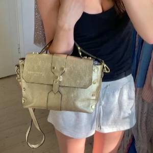 Guld axelväska från Christian Laurier - OBS knappen har trillat av (se bild 5) Snygg axelväska i metallic guld från Christian Laurier Paris. Väskan är guld glittrig med  nitar och bärhandtag samt ett justerbart axelband. Perfekt statement väska! 