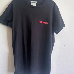 Svart Helmut Lang t-shirt med tryck - Svart t-shirt från Helmut Lang med röd text 'HELMUT' på bröstet och stort rött tryck 'IN LANG WE TRUST' på ryggen. Klassisk passform och rund halsringning. Tillverkad i mjuk bomull, perfekt för en clean och edgy look.