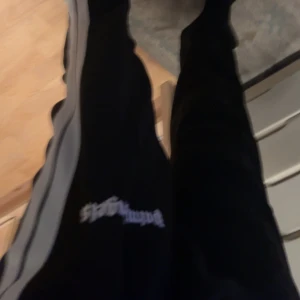 Palm Angels svarta mjukisbyxor med vita ränder - Svarta mjukisbyxor från Palm Angels med vita sidoremsor och logotyp på vänster lår. Byxorna har elastisk midja och raka ben, tillverkade i ett glansigt syntetmaterial som ger en sportig vibe. Perfekta för en chill och trendig streetwear-look.