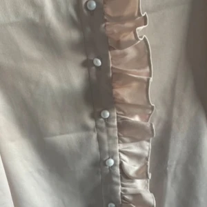 Beige blus med volang och knappar - Snygg beige blus med lång ärm, dekorativa volanger längs knäppningen och pärlemorskimrande knappar. Materialet har en lätt glans och ger en elegant känsla. Perfekt för dig som gillar feminina detaljer och stilren design.