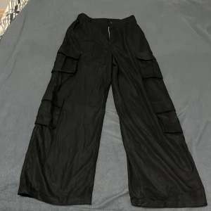 Svarta cargopants från Shein Teen - Svarta cargopants från Shein Teen med breda ben och flera stora fickor på sidorna. Byxorna har dragkedja och knapp framtill samt elastisk midja baktill för extra komfort. Perfekt för dig som gillar streetstyle och praktiska detaljer.