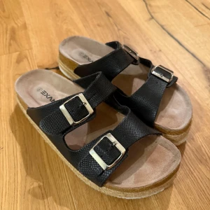 Svarta sandaler/tofflor - Snygga svarta sandaler från Exani med två breda remmar i skinnimitation och silverfärgade spännen. Plattformssula med flätad detalj ger extra höjd. Storlek 40 men passar mig som har 38/39