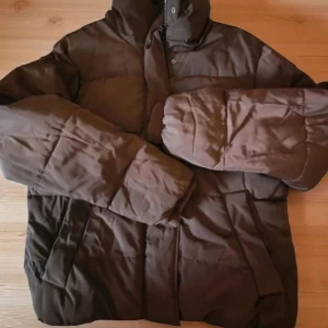 Brun pufferjacka från Dobber - Säljer en brun pufferjacka från Dobber i storlek small. Jackan har hög krage, långa ärmar och quiltad design. Perfekt för kalla dagar med sin tjocka och fluffiga känsla. Stängs med dragkedja och tryckknappar framtill.