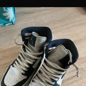 Nike Air Jordan 1 High svart/vit - Snygga Nike Air Jordan 1 High sneakers i svart och vitt skinn med klassisk swoosh på sidan. Skorna har högt skaft, snörning och rund tå. Perfekta för dig som gillar streetwear och vill sticka ut med en ikonisk modell. Något slitna på sulorna annars helt ok skick