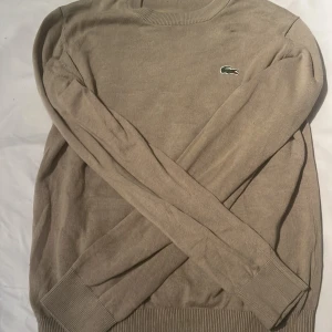 Beige tröja från Lacoste - Snygg beige tröja från Lacoste med klassisk rund hals och den ikoniska krokodilloggan broderad på bröstet. Tröjan är långärmad och tillverkad i en mjuk blandning av bomull och polyamid, perfekt för en clean och stilren look.