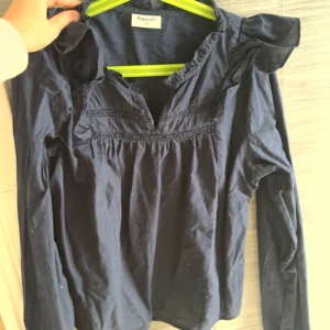 Mörkblå blus med volang från Kappahl - Säljer en mörkblå blus från Kappahl med långa ärmar och volangdetaljer vid axlar och halsringning. Blusen har v-ringning och fina rynkade partier framtill. Perfekt för dig som gillar en söt och stilren look.