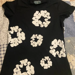 Svart blommig t-shirt från Denim Tears - Svart t-shirt från Denim Tears med coola vita blommönster tryckta i cirklar över hela framsidan. Klassisk rund hals och korta ärmar. Materialet är mjuk bomull som känns skönt mot huden. Perfekt för dig som gillar streetwear med unik design.