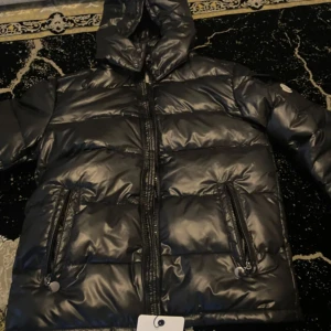 Svart Moncler pufferjacka - Säljer en svart pufferjacka från Moncler med glansig finish och huva. Jackan har två fickor med dragkedja framtill och klassisk Moncler-logga på ärmen. Perfekt för kalla dagar och riktigt snygg streetstil.