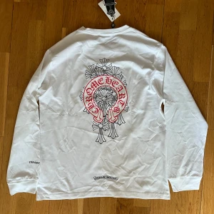 Vit långärmad tröja Chrome Hearts - Vit långärmad tröja från Chrome Hearts med rött tryck på bröstet och stor grafisk design på ryggen. Tröjan har rund halsringning och är tillverkad i mjuk bomull. Snygga detaljer med logga på ärmen och nertill på ryggen. Perfekt för dig som gillar streetwear vibes. Storlek L. Säljer pga för liten. 