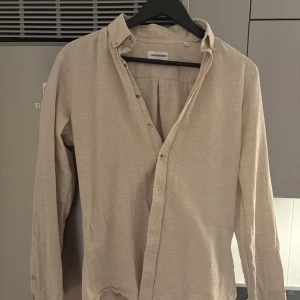 Beige linneskjorta från Lindbergh - Snygg beige skjorta från Lindbergh i mjukt linne. Klassisk krage, knappar framtill och långa ärmar. Perfekt för dig som gillar en clean och stilren look. Skjortan har en avslappnad passform och passar till många olika outfits.